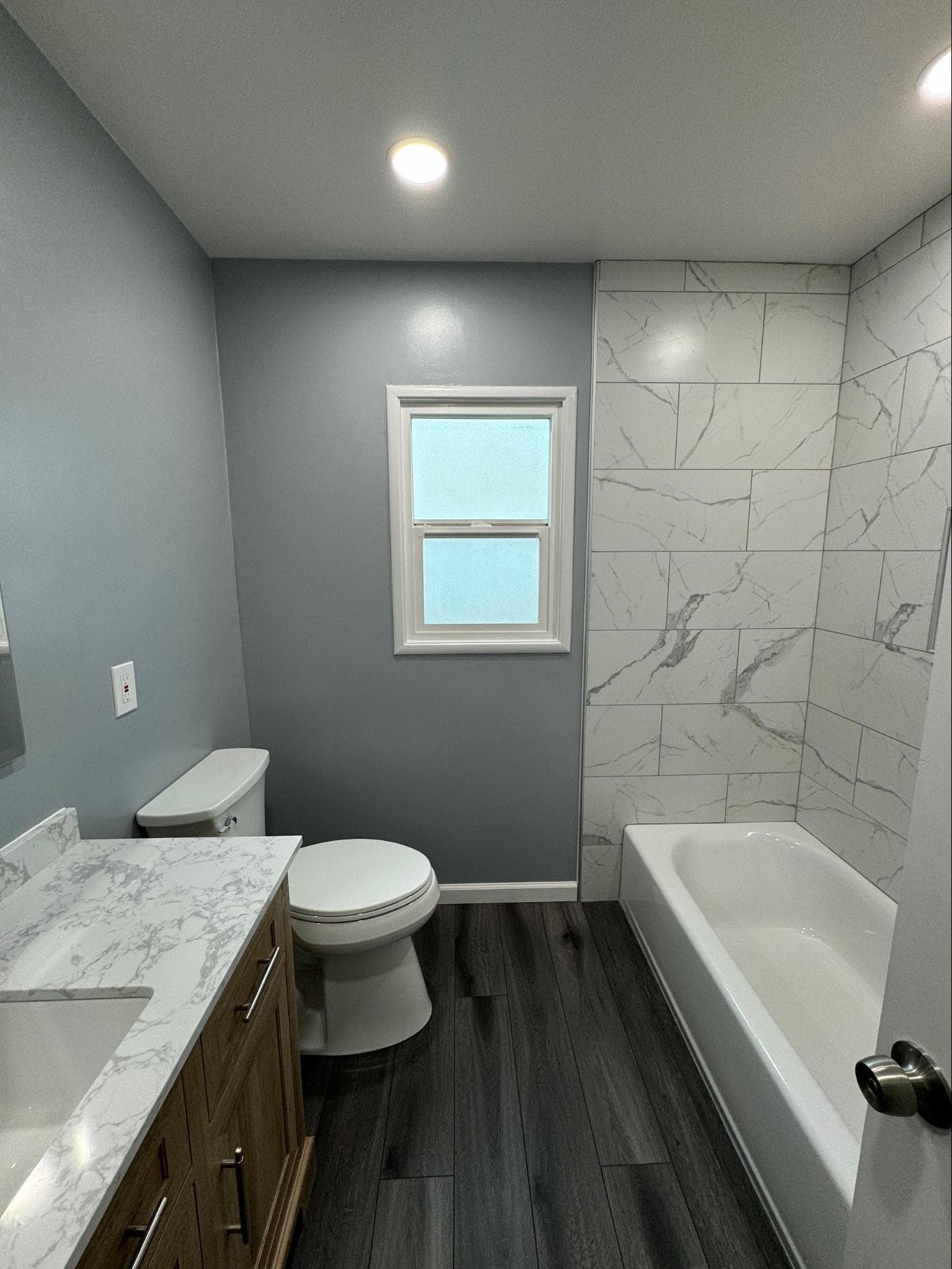 Modern bathroom featuring gray walls, marble-tiled shower, white toilet, and dark wood-look flooring.